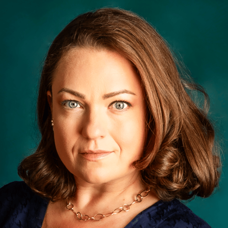 Headshot of a smiling woman with brown hair, blue eyes, and a gold necklace.