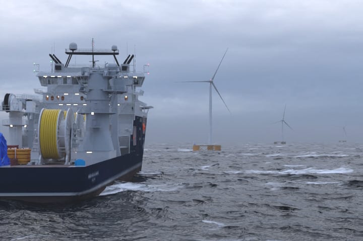An offshore service vessel with lights on approaches a floating wind turbine in darkening weather conditions.