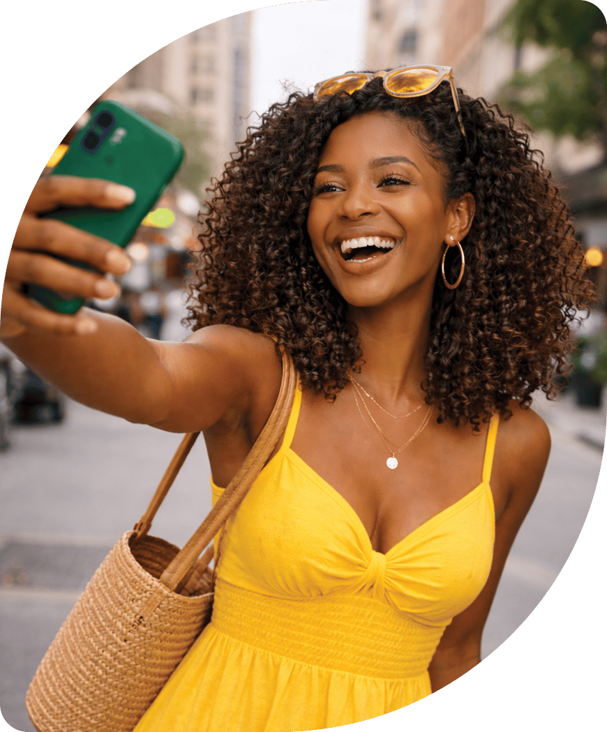 A joyful woman with curly hair in a yellow dress smiles while taking a selfie with her green phone.