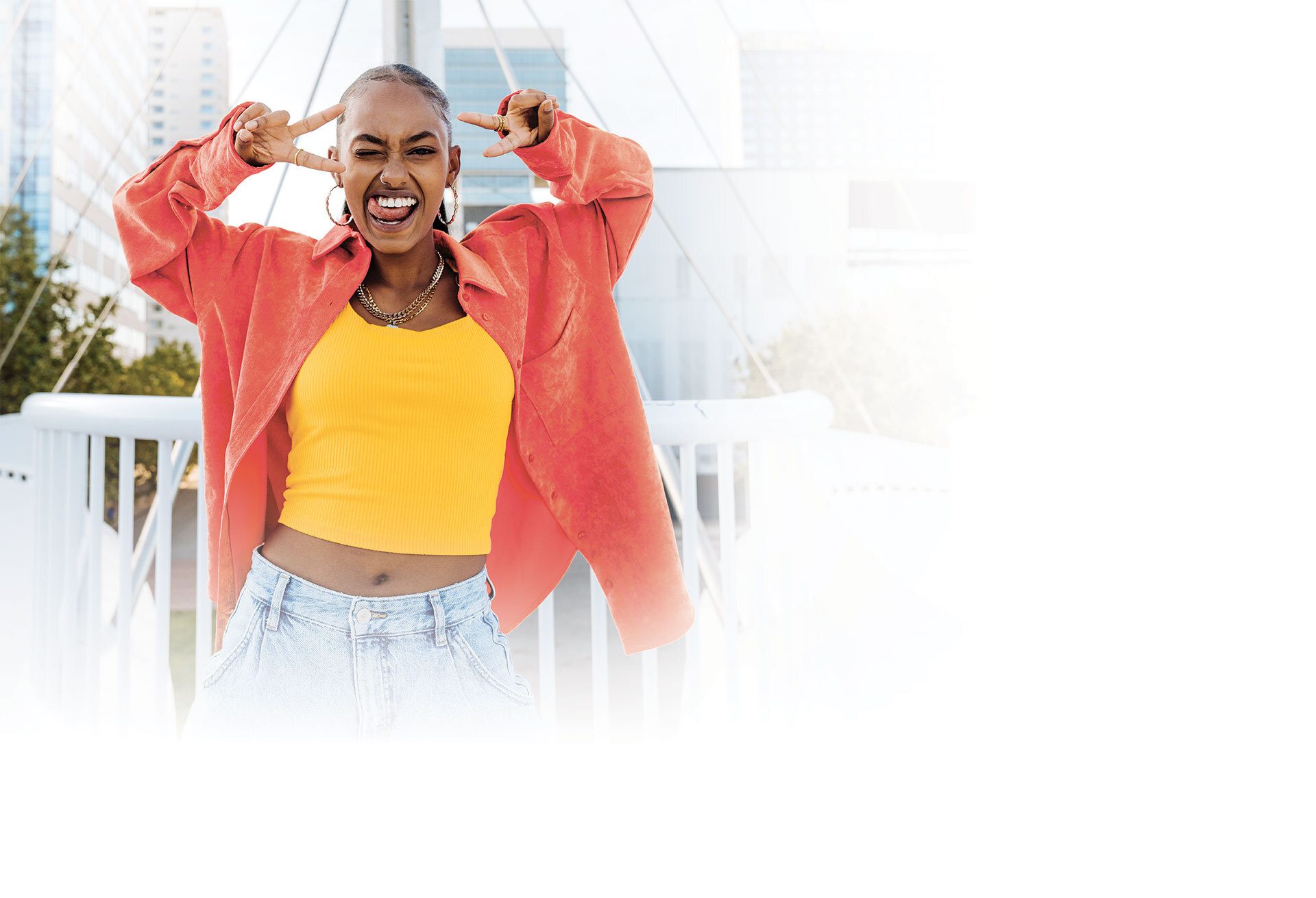 Woman winking, tongue out, making peace signs in a coral jacket and yellow top on a bridge.
