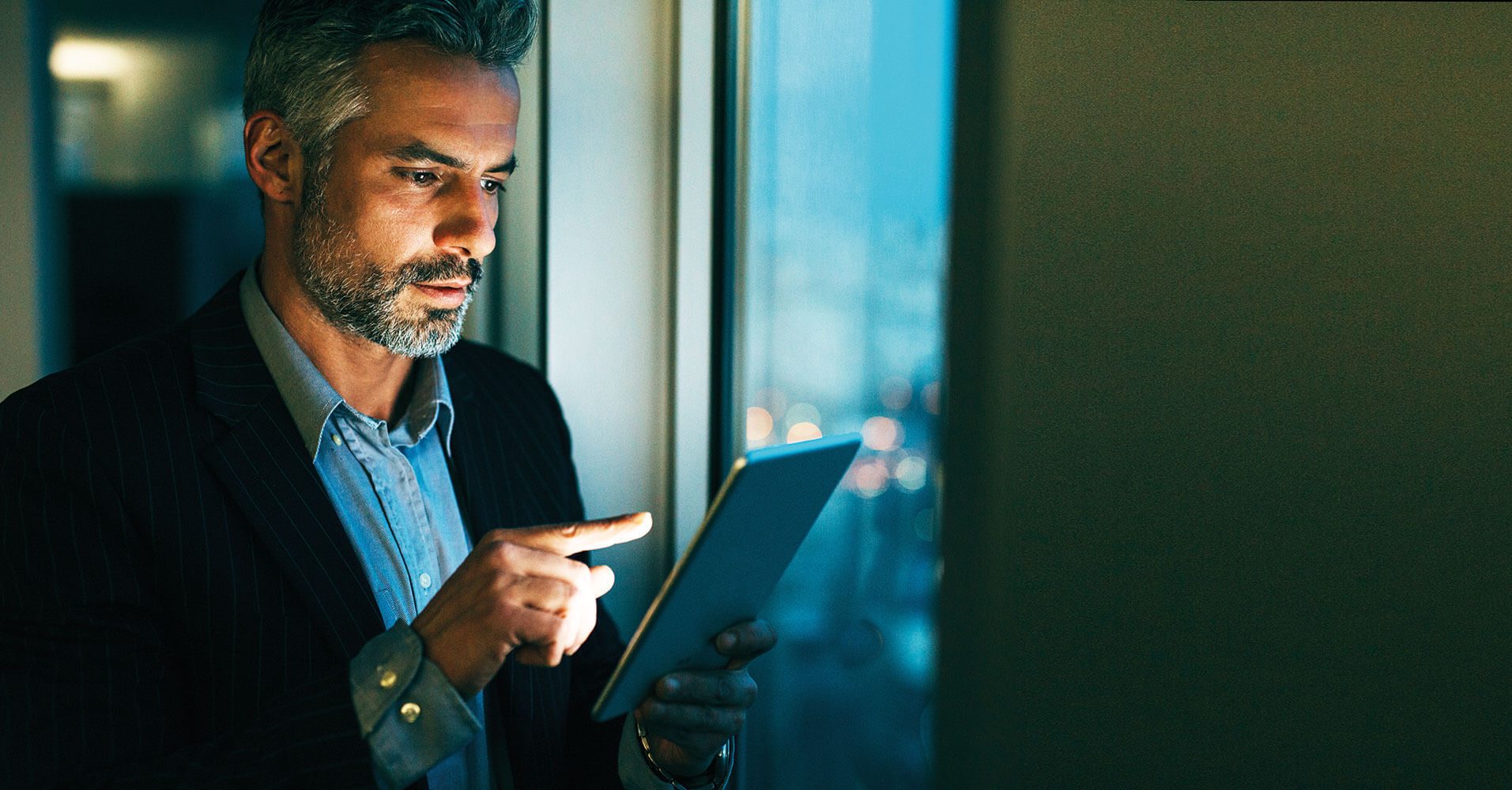 A focused businessman uses a tablet, illuminated by its screen and window light, in a dark setting.