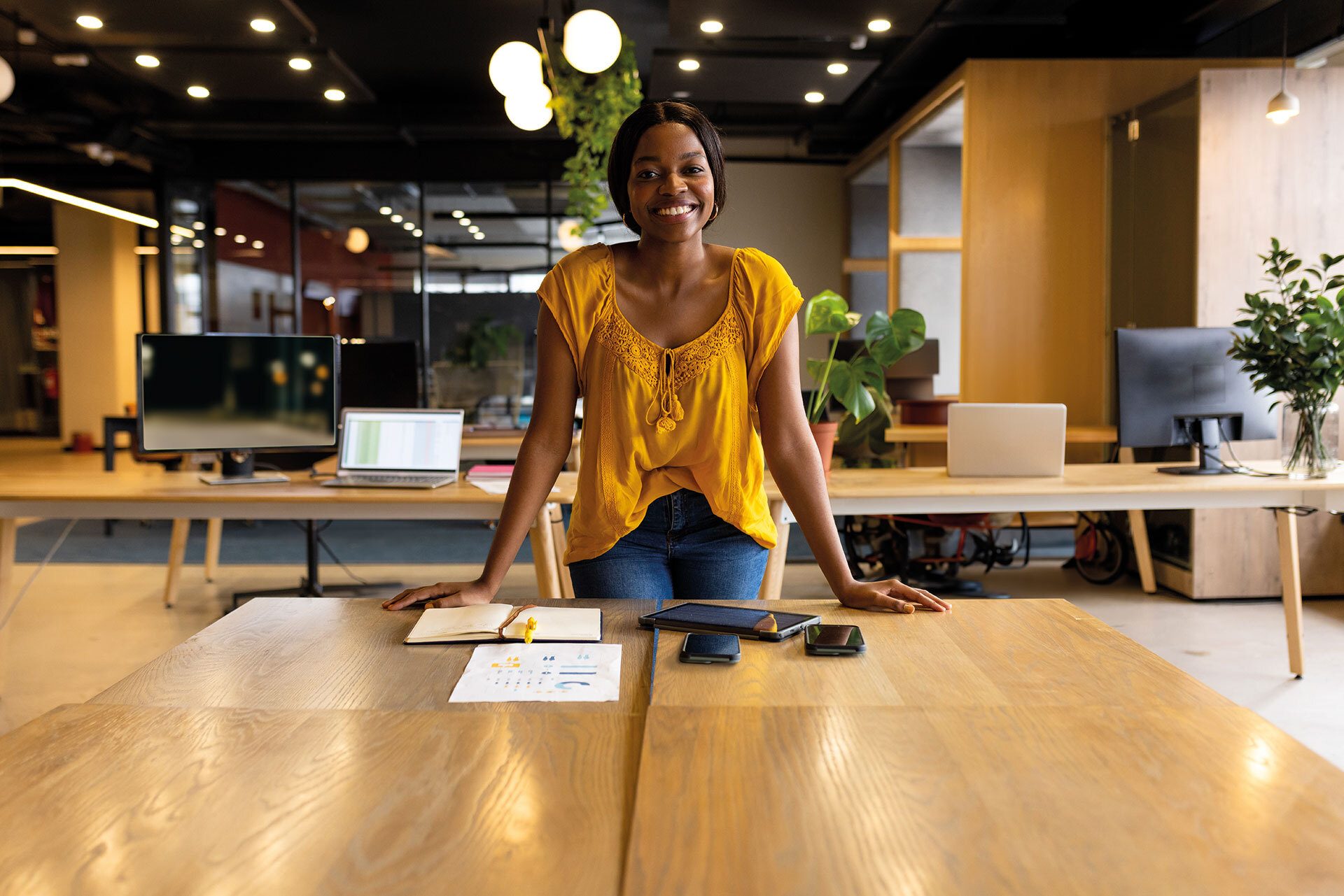 Smiling Black woman in a yellow top and jeans at a wooden office table with tech and papers.