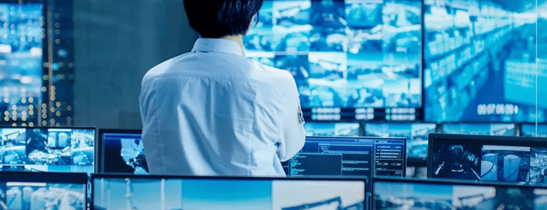 A security guard monitors multiple screens displaying surveillance footage and data in a control room.