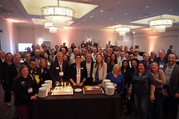 Large group of people celebrating around two cakes, one with "75" candles, at an event.