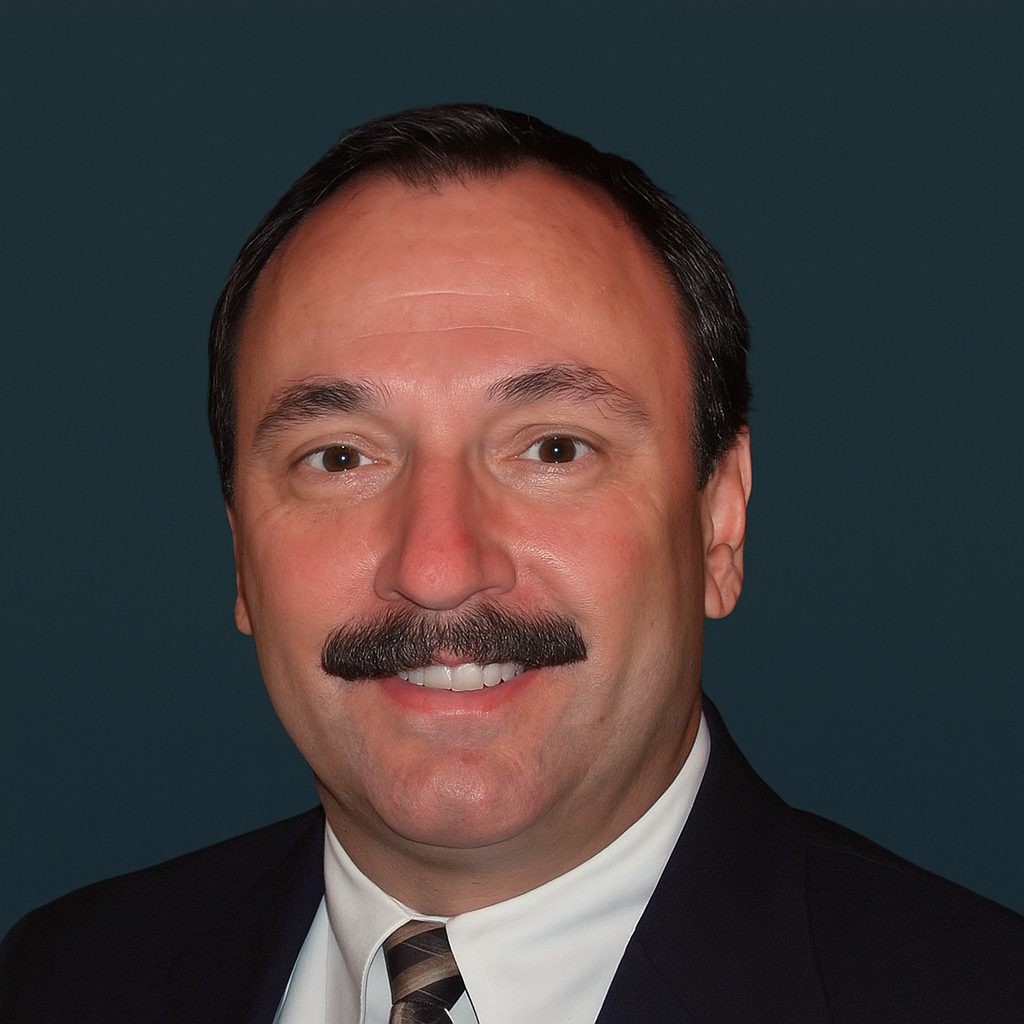 Smiling man with dark hair and a mustache, wearing a suit and tie, against a dark background.
