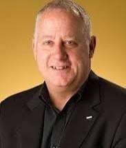 Smiling man with short gray hair in a black suit against a yellow background.