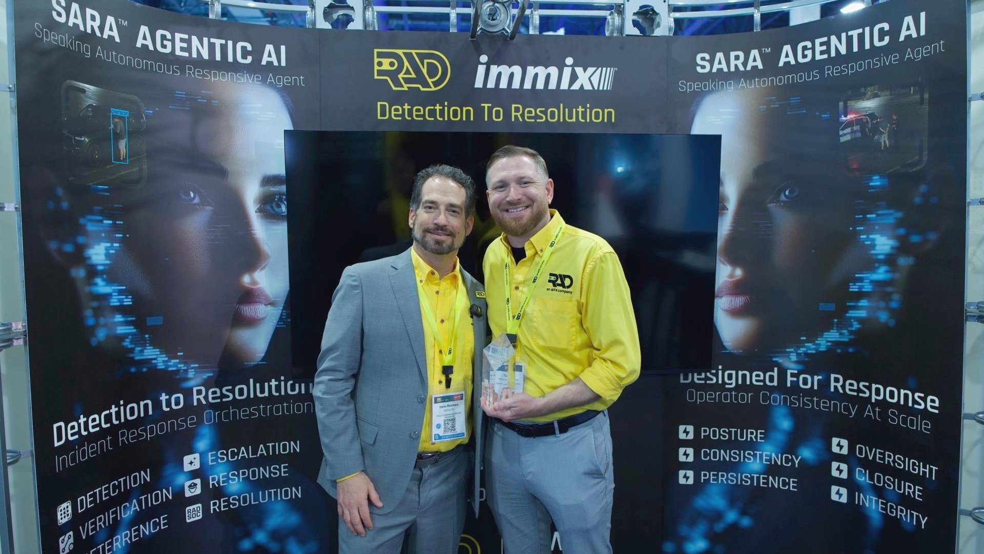 Two men smiling at an AI security booth with "SARA Agentic AI" and "RAD immix Detection To Resolution".