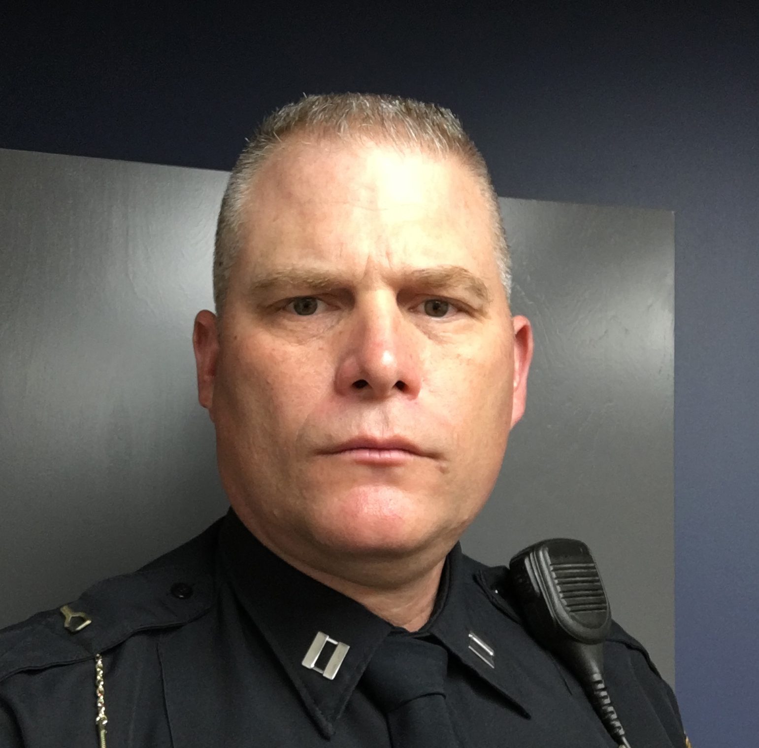 Headshot of a serious man in a law enforcement uniform, with a shoulder radio.