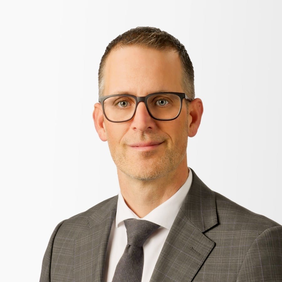 Headshot of a man in a gray suit, white shirt, and gray tie, wearing dark-framed glasses.