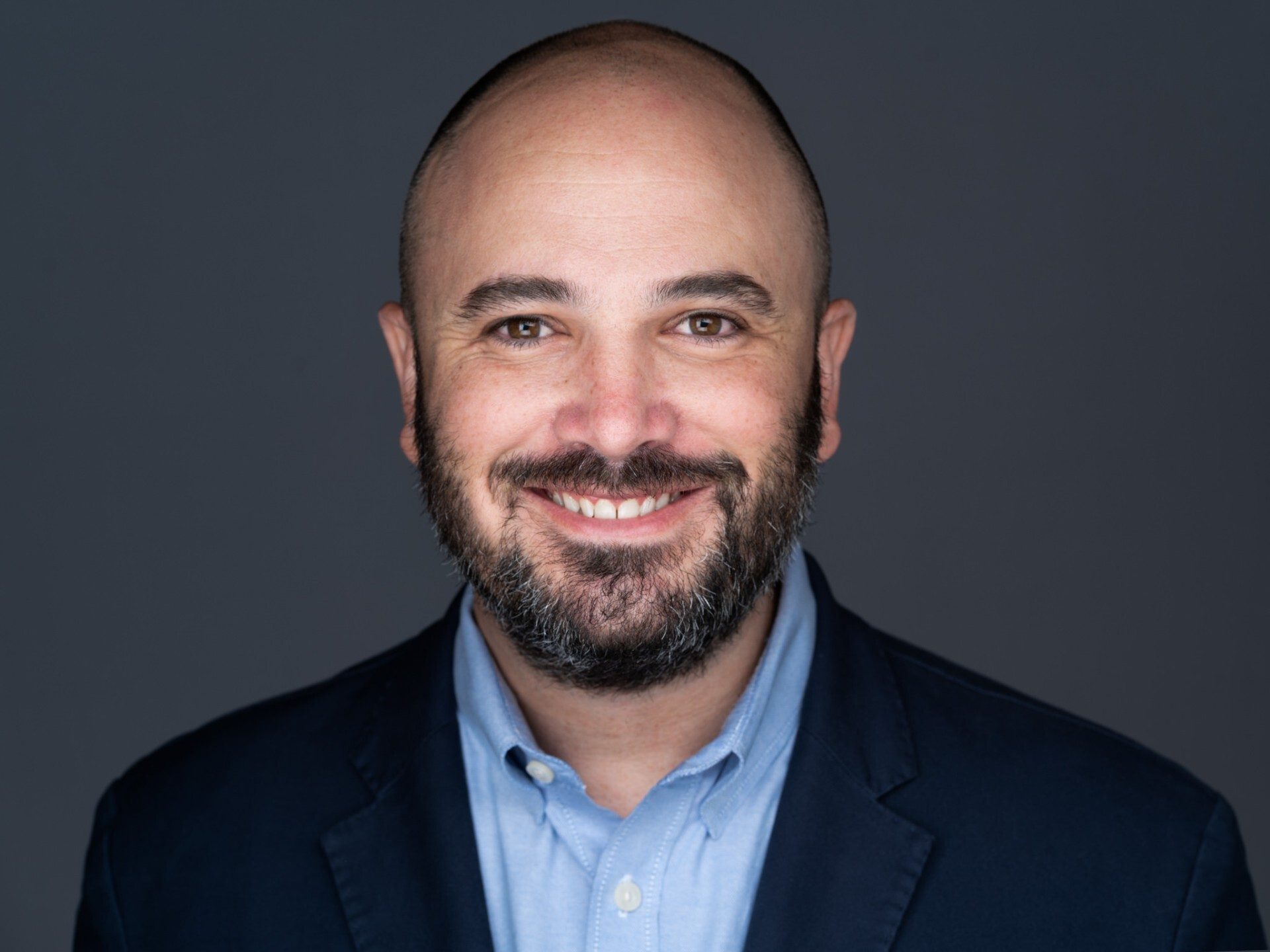 Headshot of a smiling bald man with a beard, wearing a blue shirt and dark blazer.