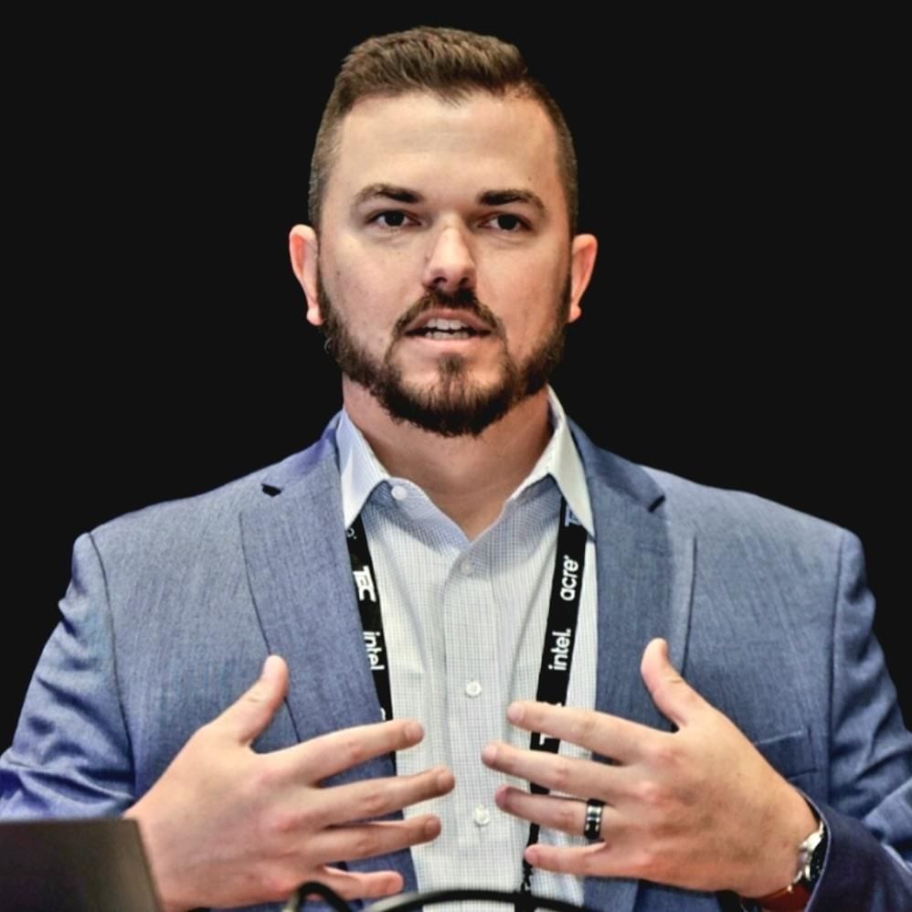 Man in blue suit jacket speaking, wearing Intel lanyards, with a beard and hands raised.