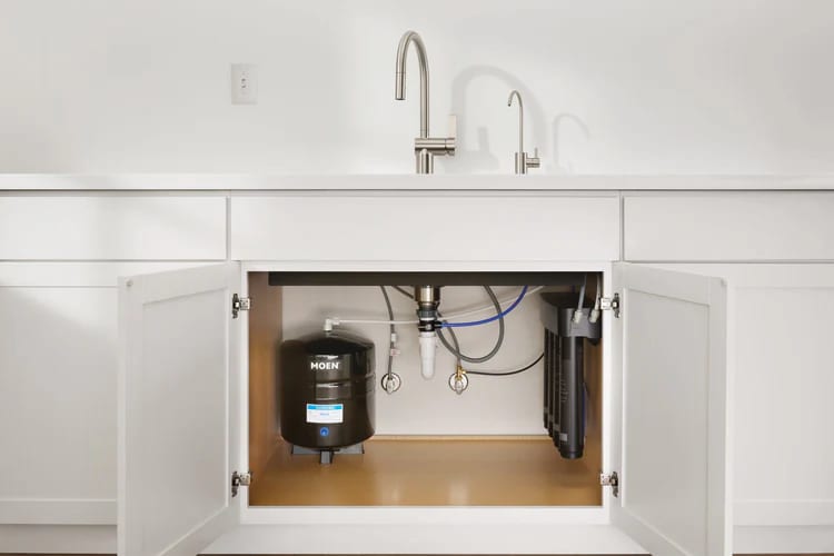 Under-sink water filtration system with tank, filter, sink, and two faucets in a white kitchen.