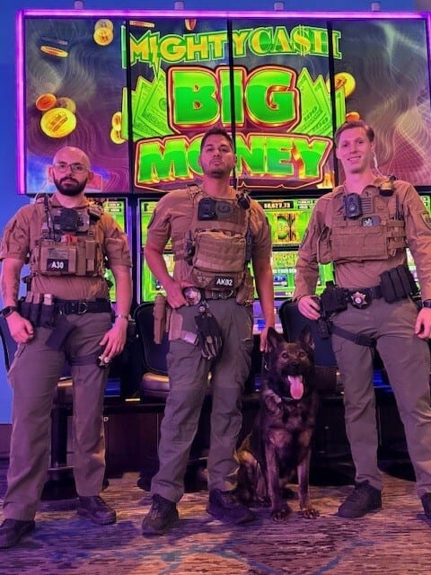 Three officers and a K9 dog stand in front of slot machines that say "MIGHTY CASH BIG MONEY".