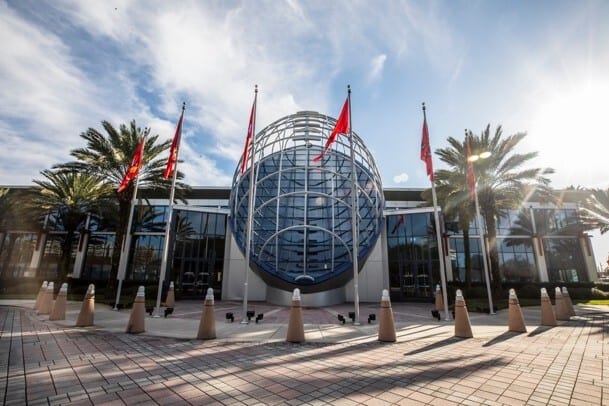 Modern building with a large spherical glass structure, red flags, palm trees, and a paved entrance.
