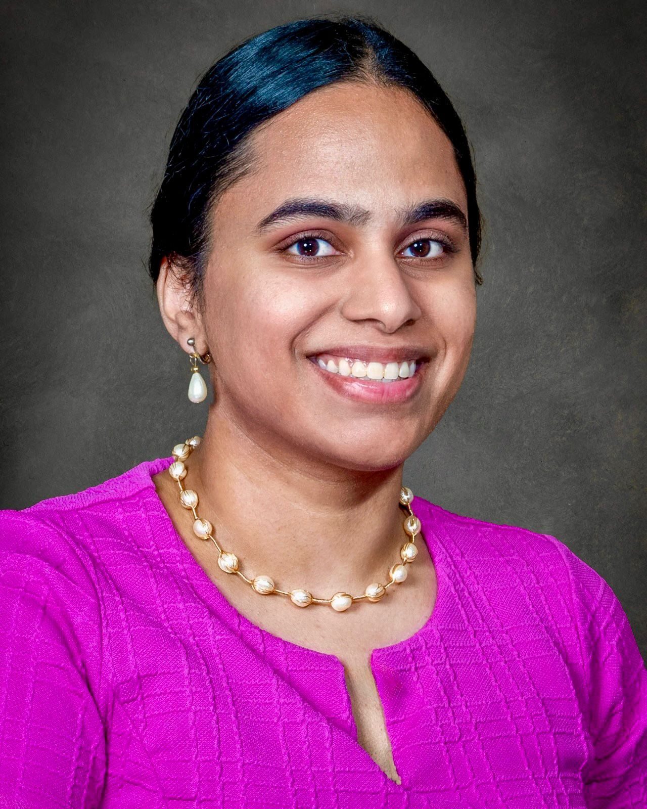 Smiling woman with dark hair, wearing a magenta top, pearl necklace and earrings.