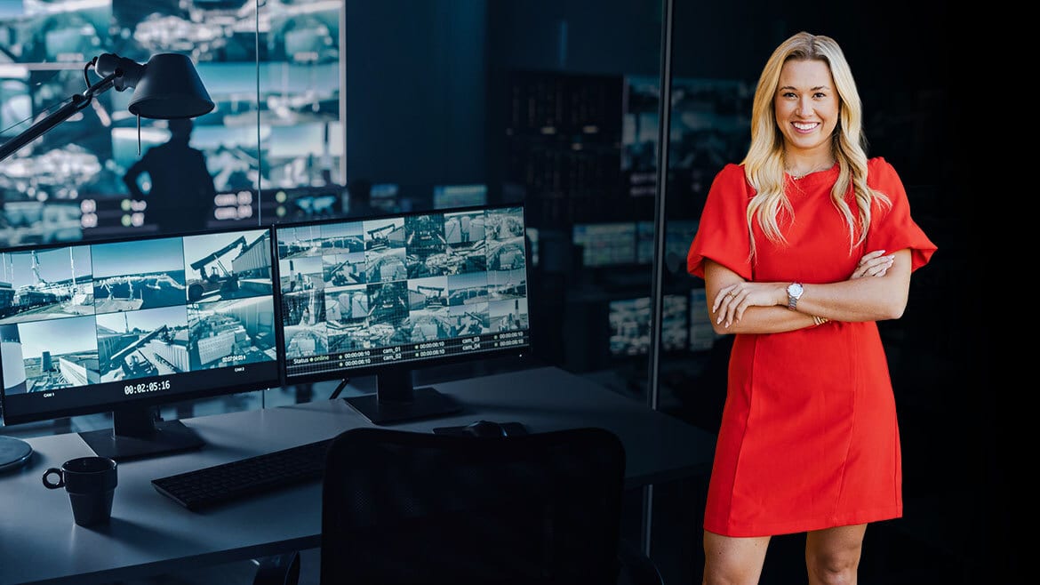 Smiling woman in red dress stands beside a desk with multiple monitors displaying security camera feeds.