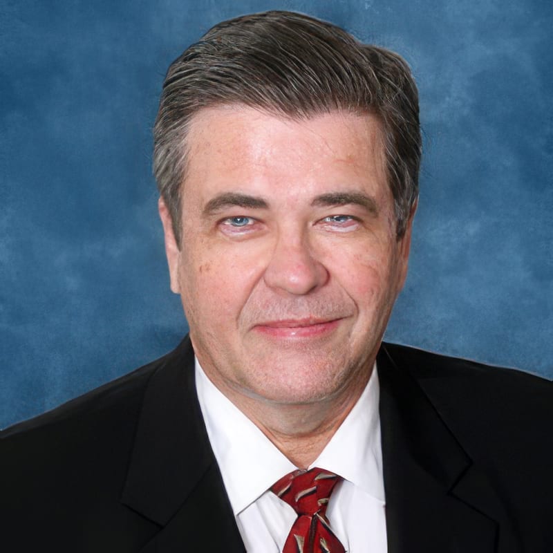 A headshot of a middle-aged man with gray hair, blue eyes, wearing a black suit and a red tie.