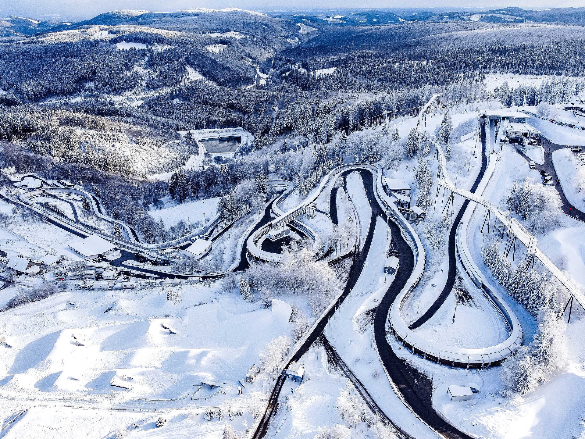 Aerial view of a winding, snow-covered bobsled track in a winter mountain forest.
