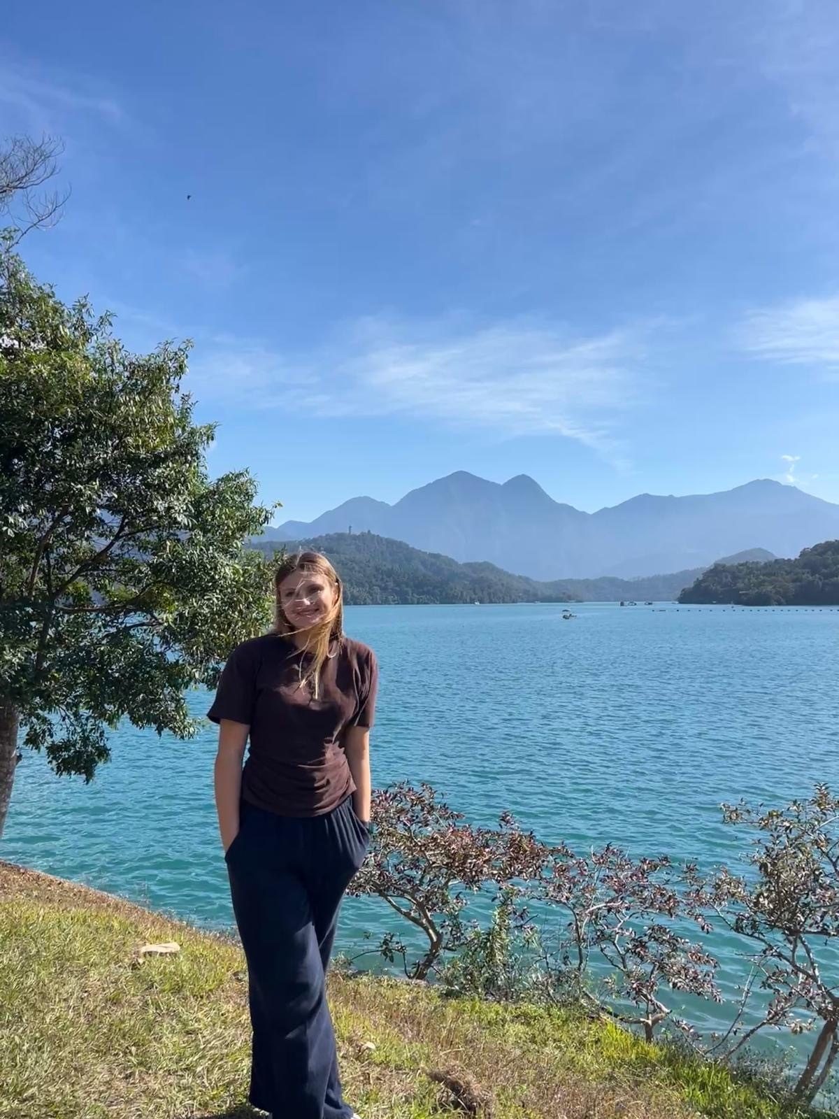 A young woman stands by a bright blue lake with mountains in the background under a clear sky.