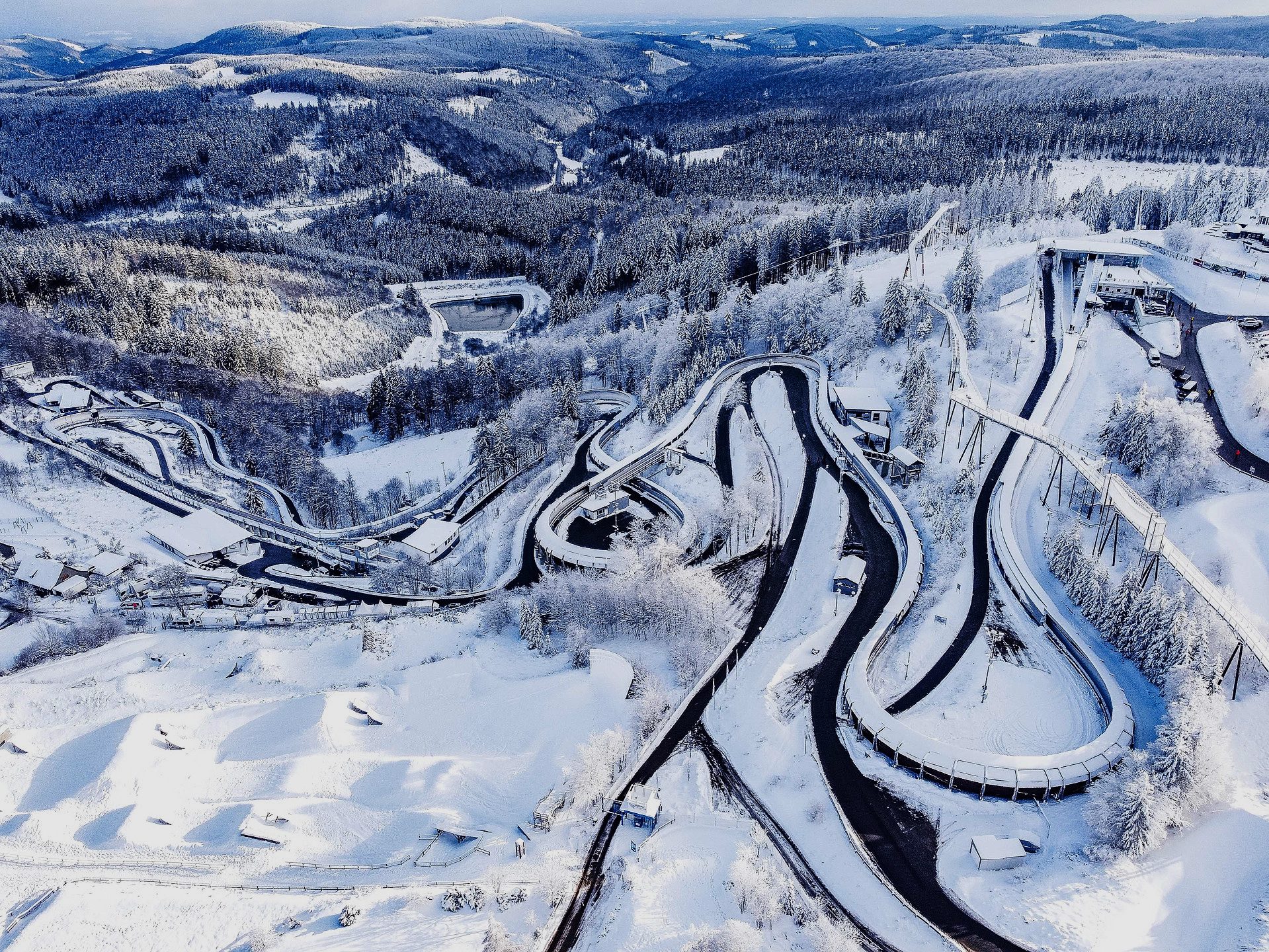 Aerial of a winding, snowy bobsled track snaking through forested mountains in winter.