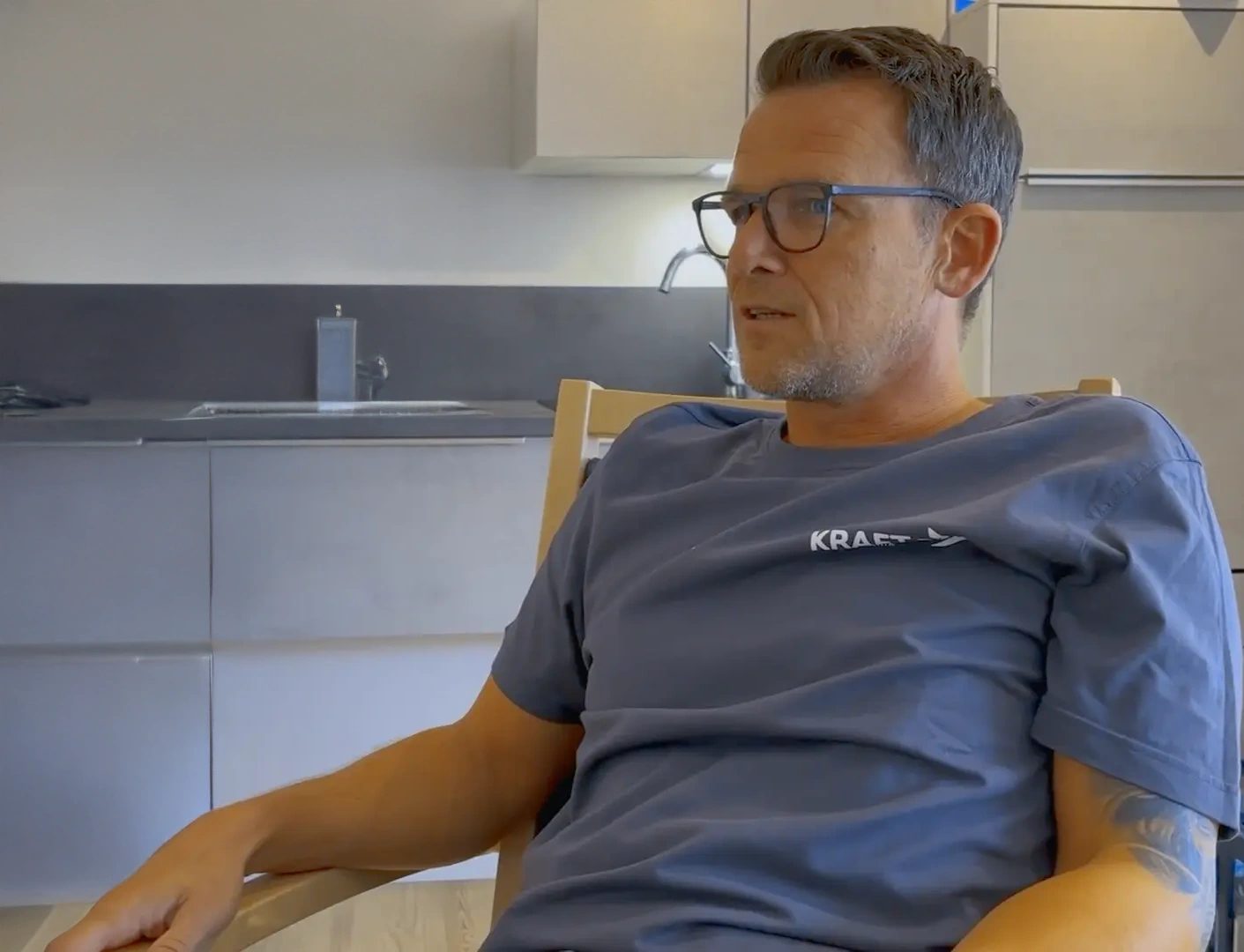 Man with glasses and a blue t-shirt, sitting indoors, likely in a modern kitchen setting.