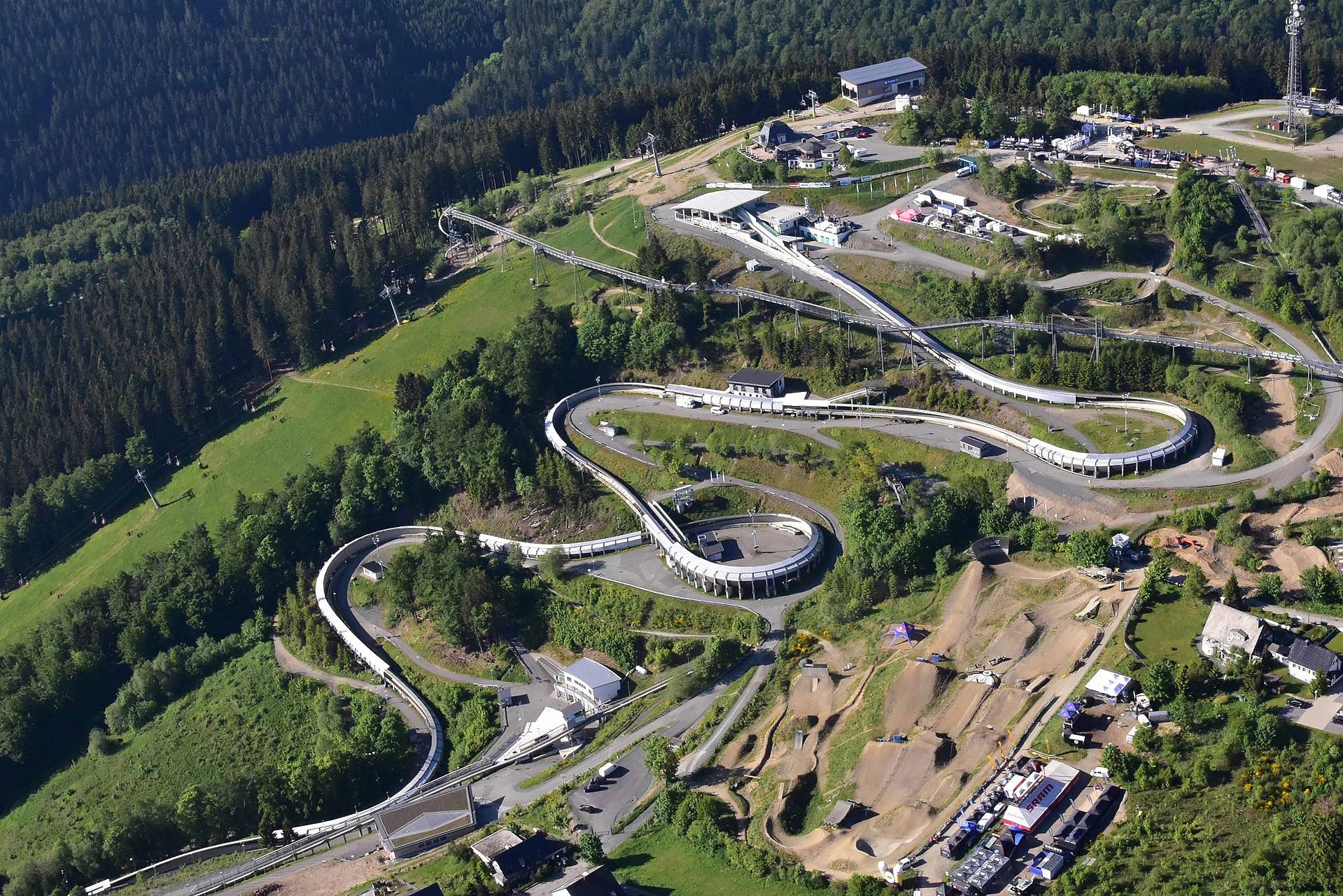 Aerial view of a winding bobsled track and mountain bike park on a lush, green forested mountain.
