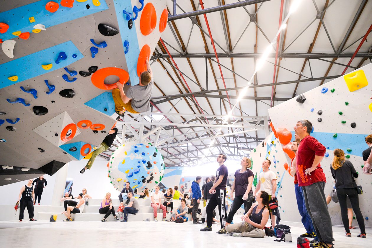 Indoor bouldering gym with climbers on colorful walls, and people resting and watching.
