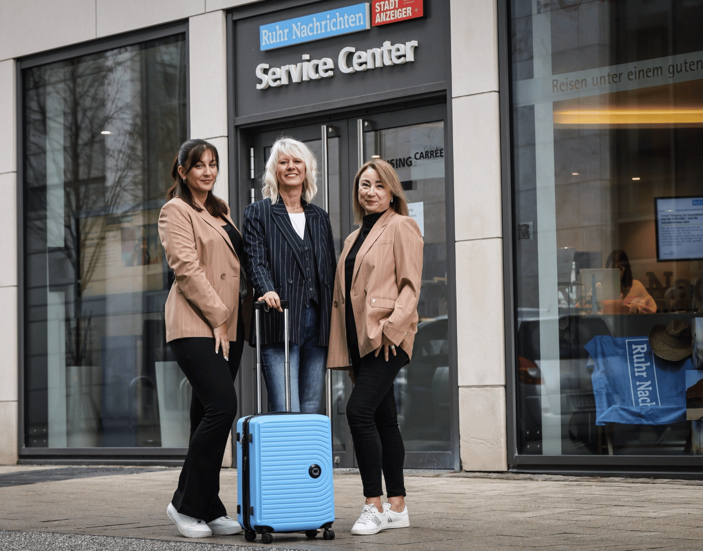 Three women stand outside a "Ruhr Nachrichten Service Center" with a blue suitcase.