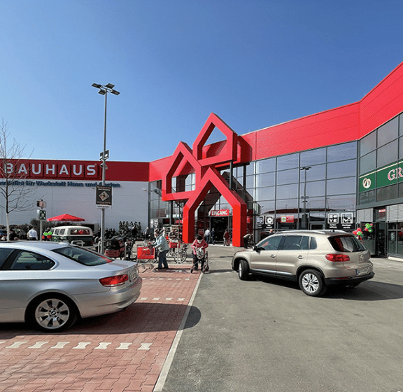 Red Bauhaus store with a distinctive geometric entrance, cars, and people in a parking lot.