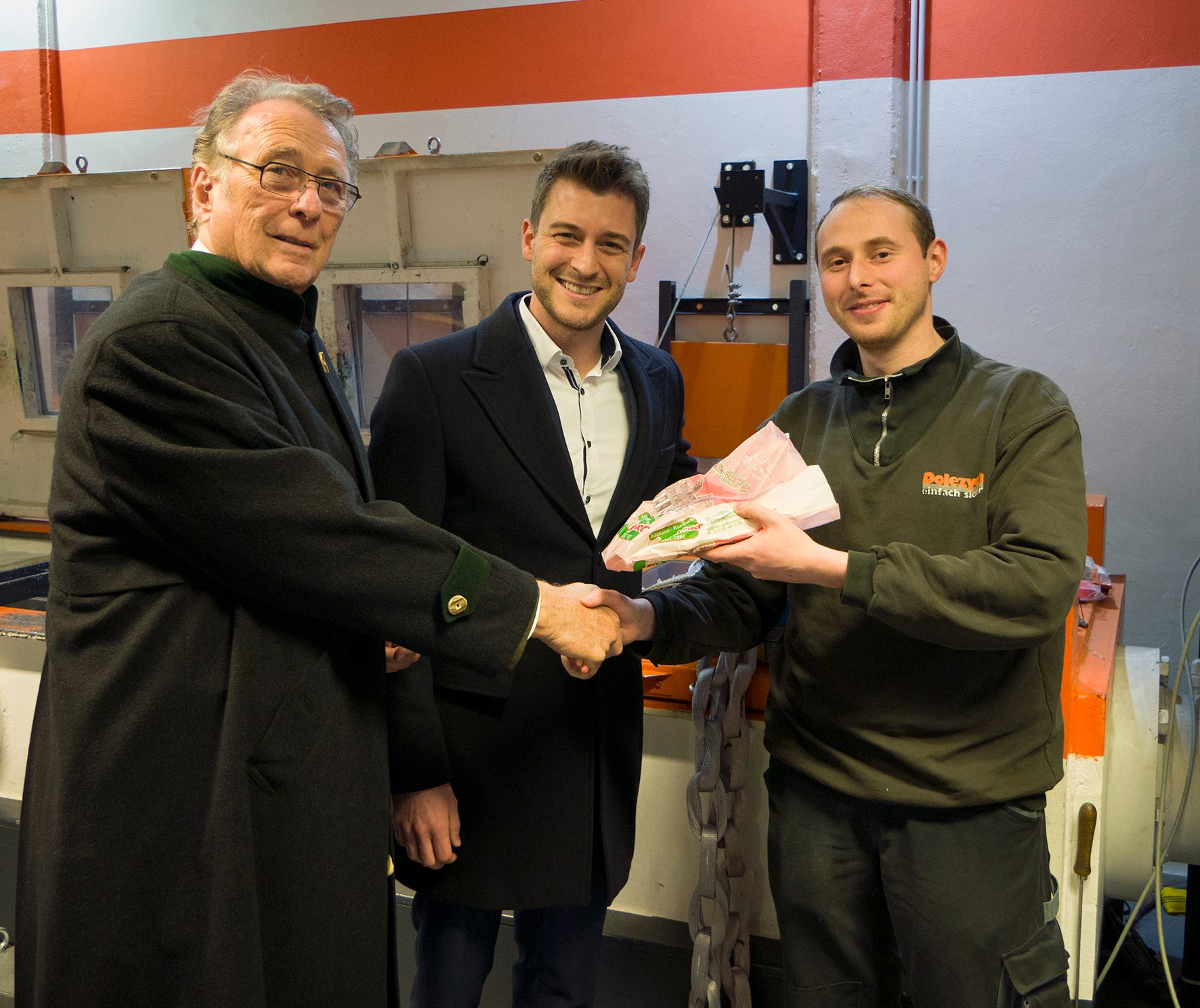 Three men linked by handshakes in a workshop; the rightmost man holds packages.