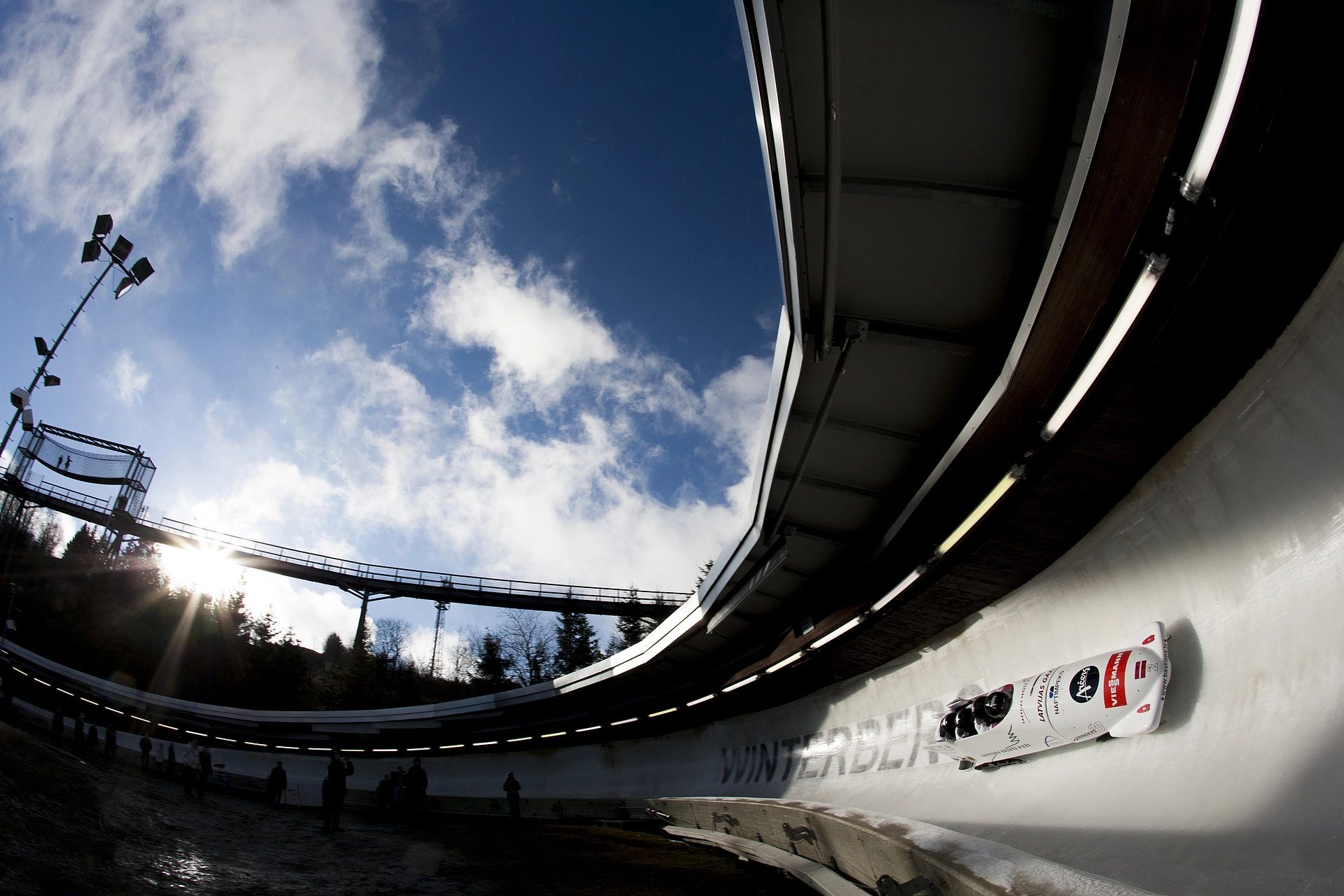 A white bobsled on a high-banked icy track under a blue sky with a sunburst.