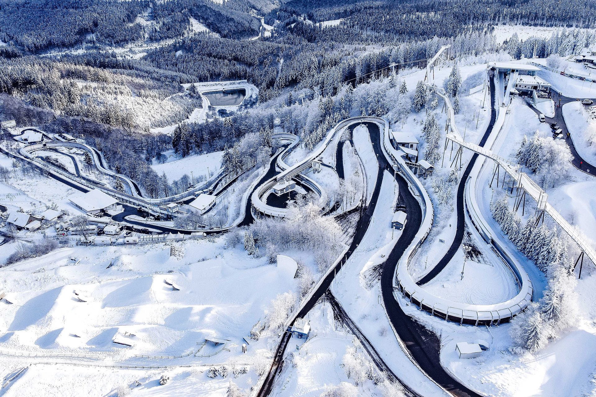 Aerial view of a winding bobsleigh/luge track and ski jumps in a snowy mountain forest.