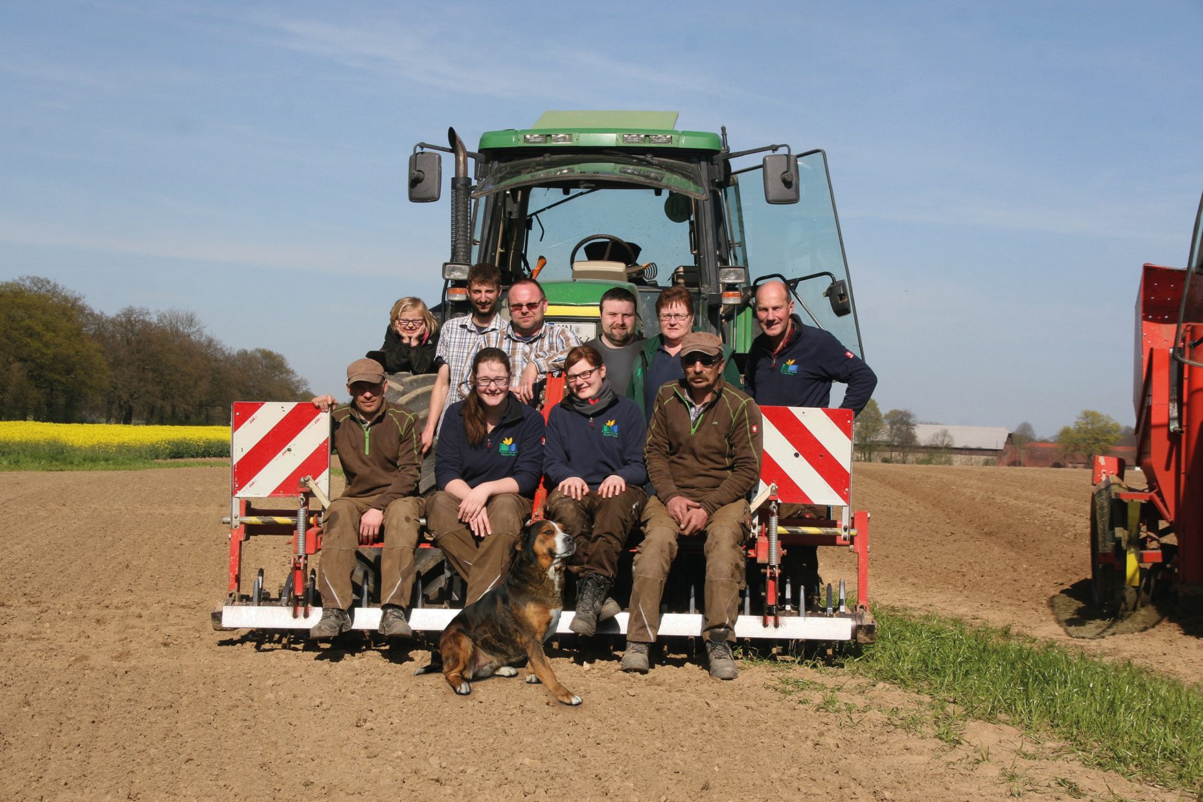 Group of people and a dog by a tractor and farm equipment in a field with yellow flowers.