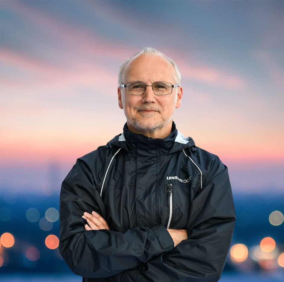 Smiling man in glasses and dark jacket, arms crossed, against blurred sunset city lights.