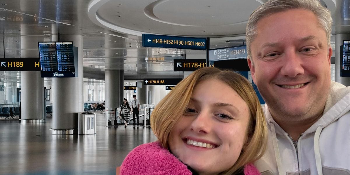 A man and a young woman smile, taking a selfie in a brightly lit airport terminal with gate signs visible.