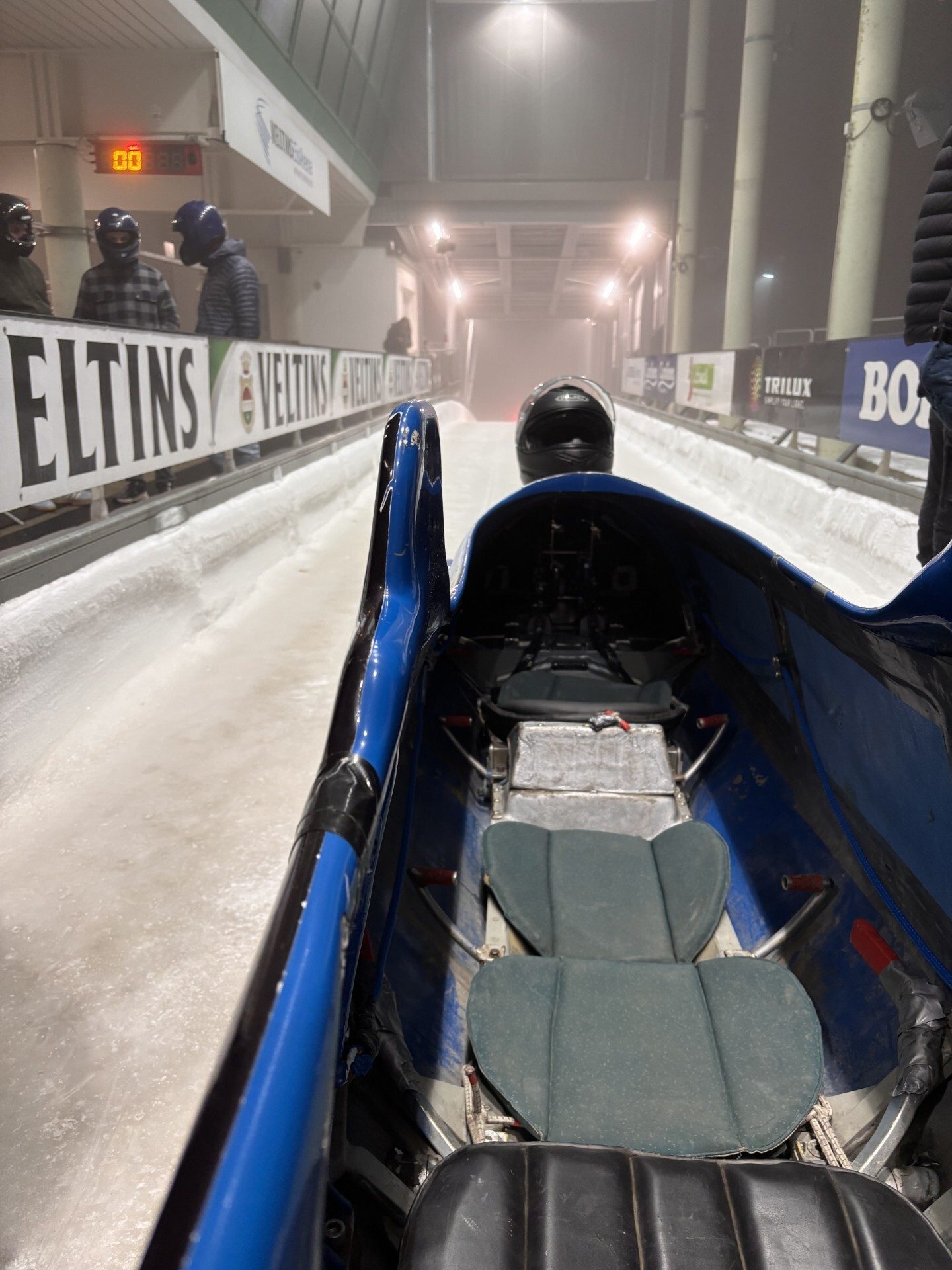 View from inside a bobsled on an icy track, with people, a timer, and sponsor signs.