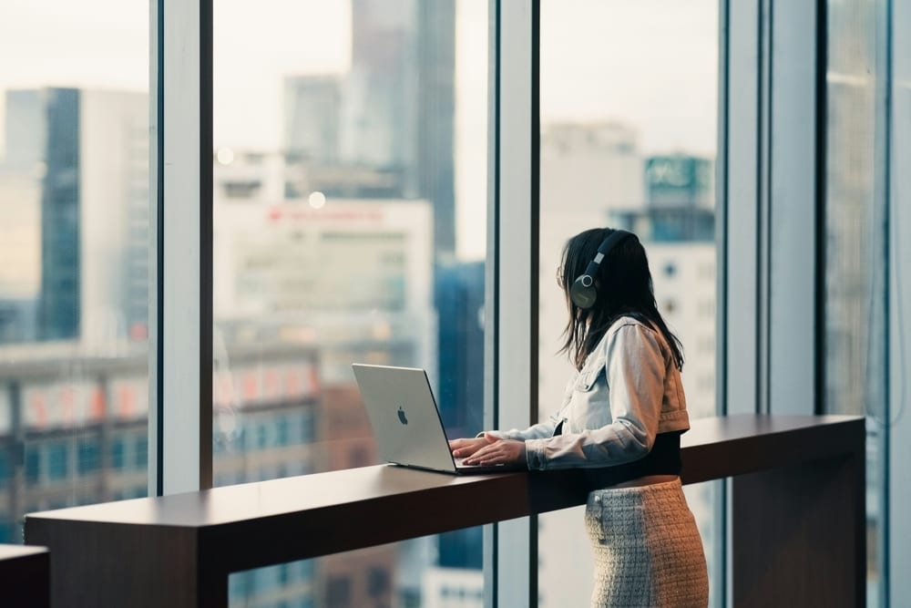 Person in headphones works on laptop by window overlooking a city.
