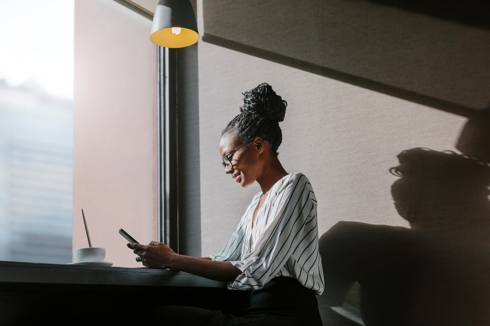 Smiling Black woman with glasses and braided bun, looking at her phone by a window, strong shadows.