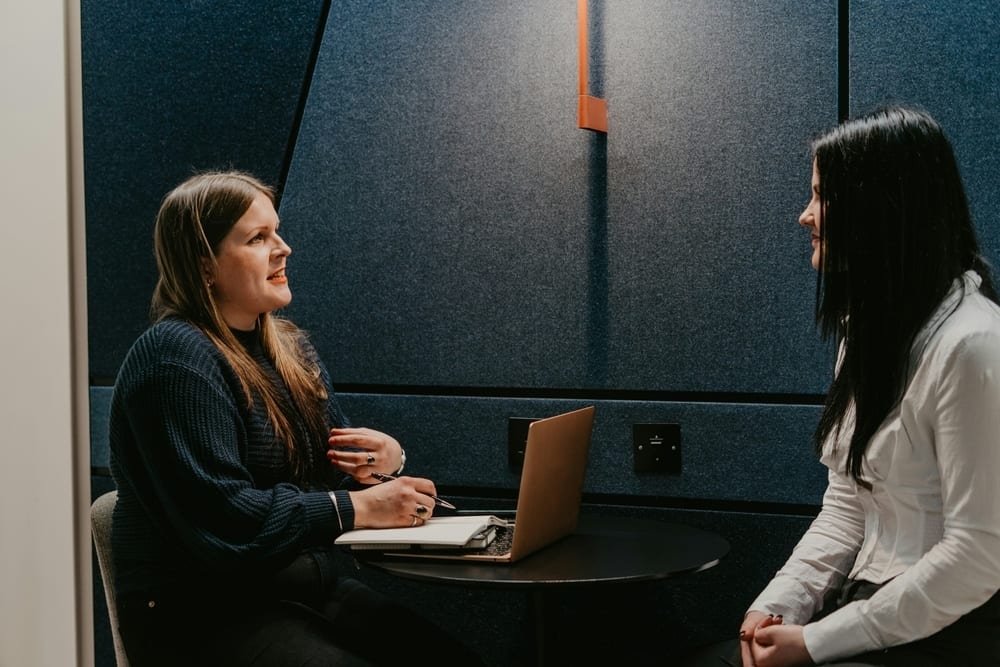 Two women conversing in a modern office nook, one taking notes next to a laptop.