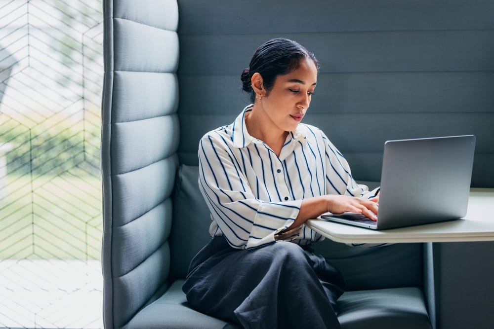 Woman working on laptop in a cozy booth by a window.
