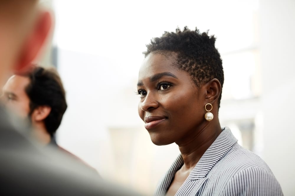 Smiling Black woman with short textured hair, pearl earrings, and striped blazer, looking right.