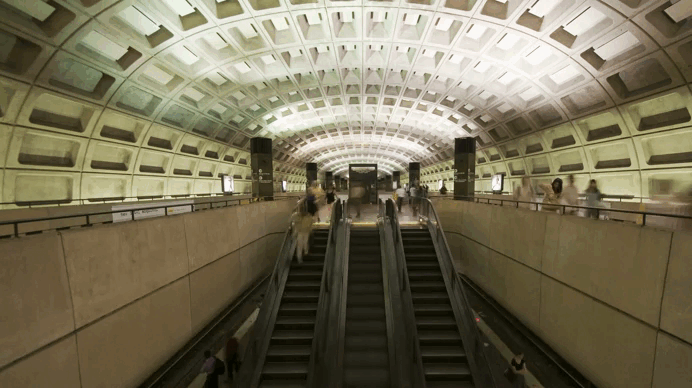 Subway station with an arched, coffered ceiling, escalators, and blurred commuters.