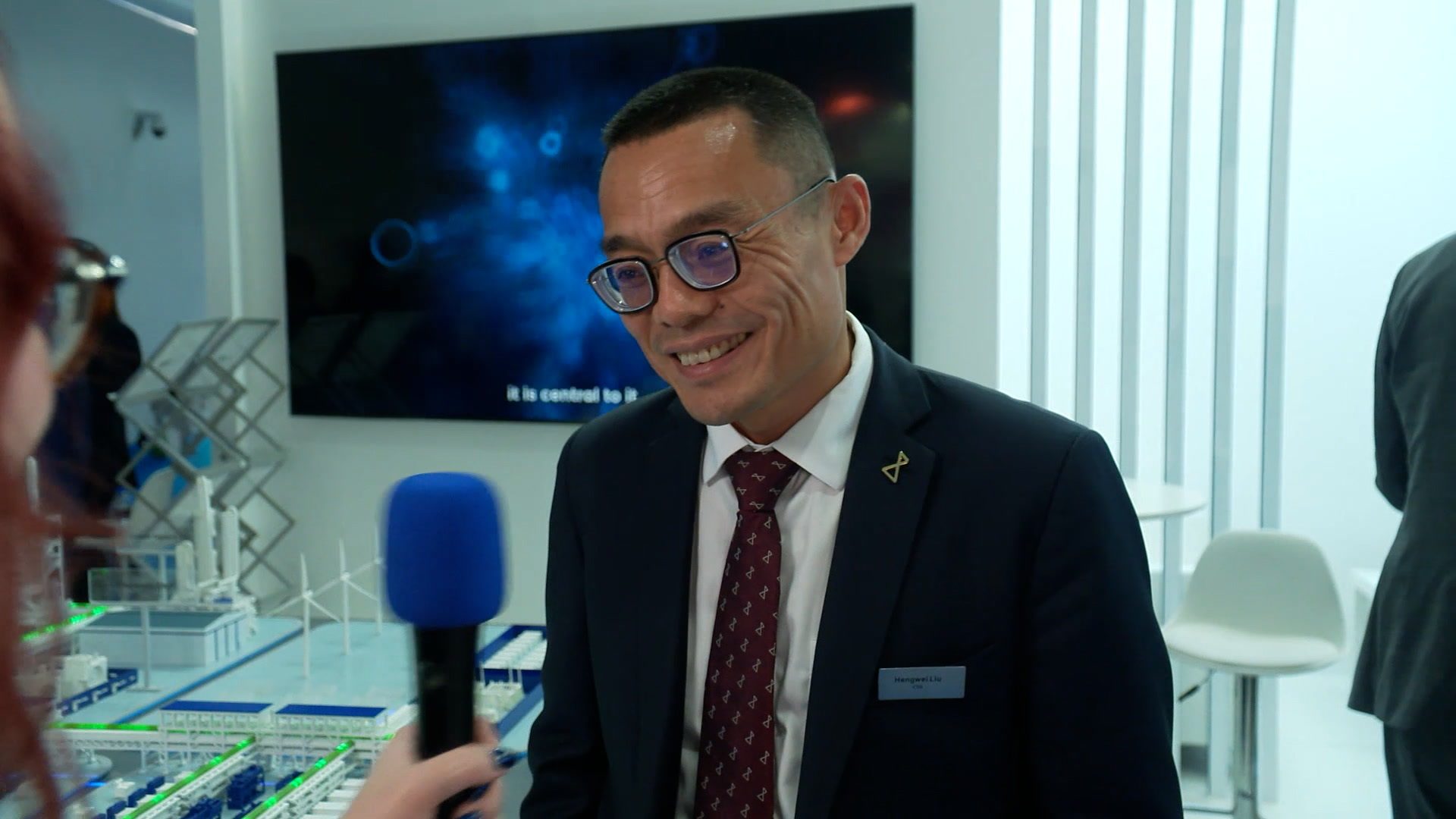 Smiling man (Hengwei Liu) in glasses and suit interviewed with microphone at an exhibition booth.
