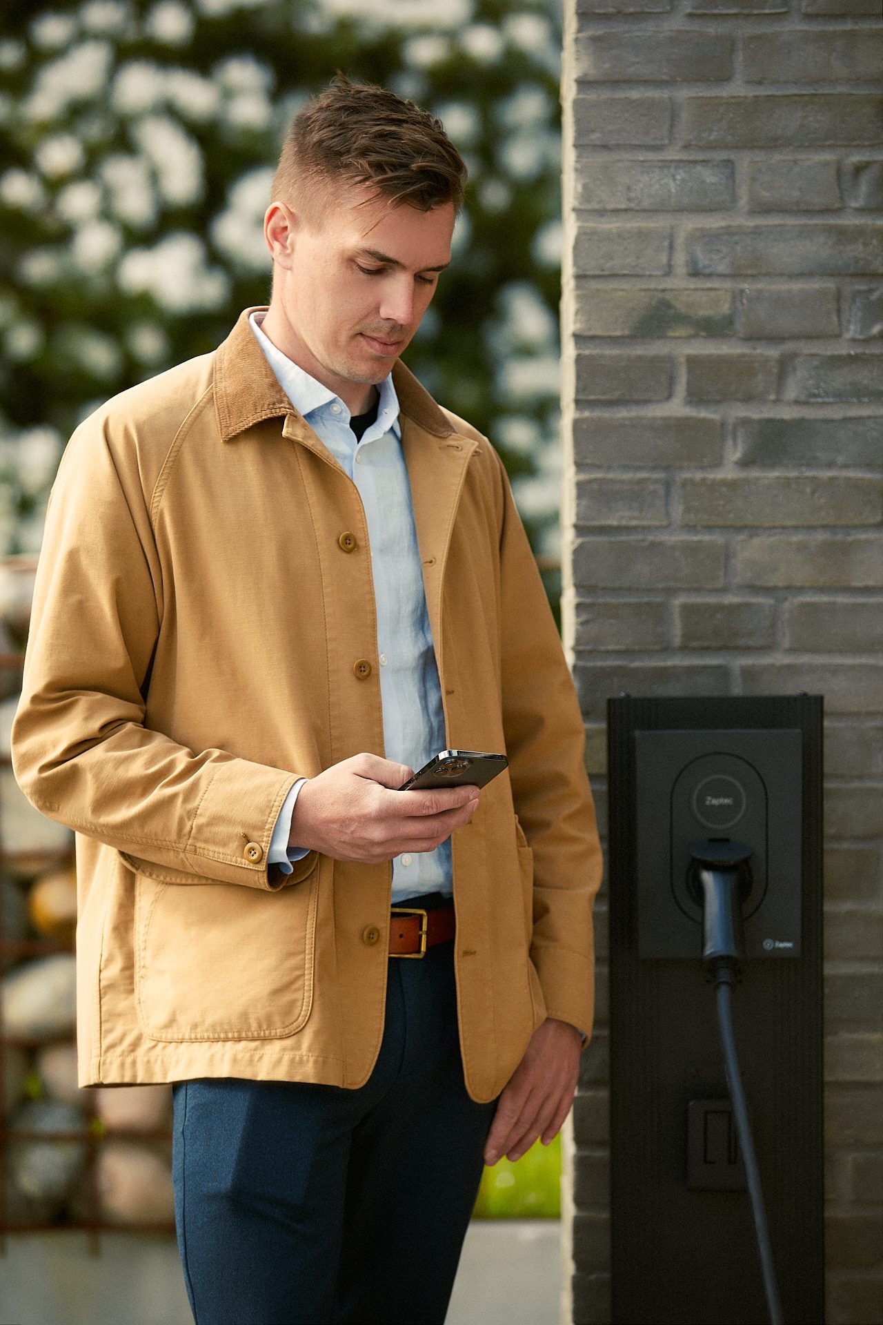 Man in brown jacket checking his phone next to an electric vehicle charging station.