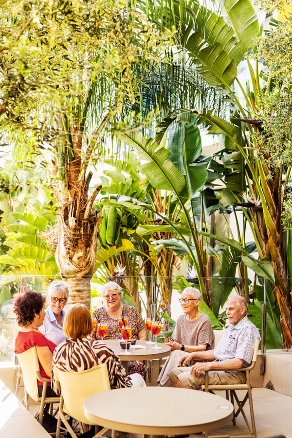 Six seniors enjoying drinks at an outdoor cafe surrounded by lush tropical plants.