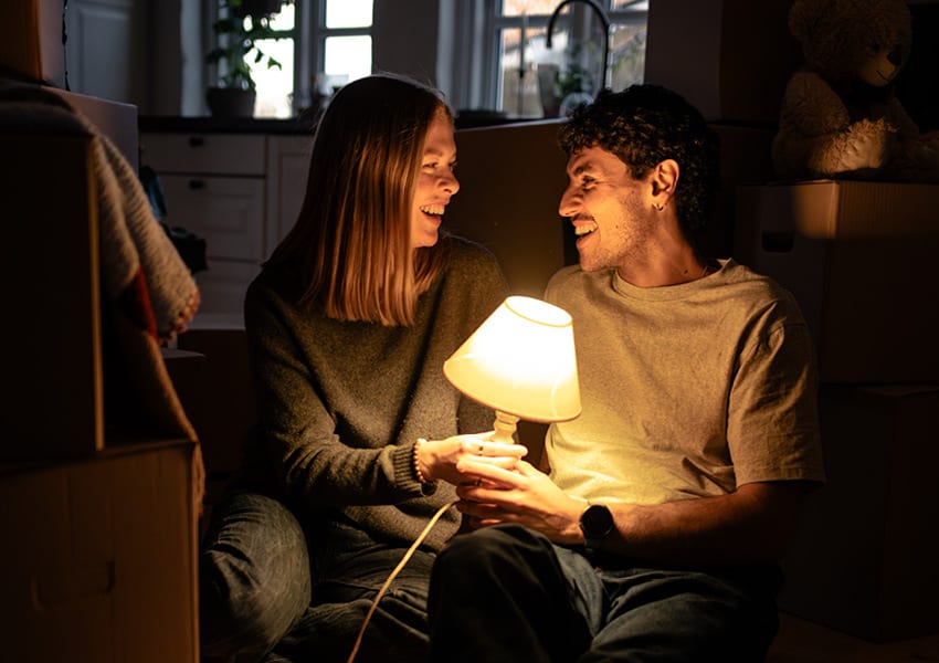 Couple sitting in the dark with a lamp, laughing amidst moving boxes.