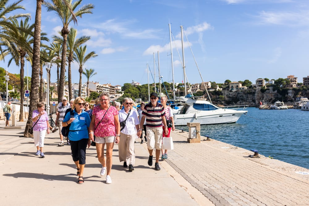 A group of seniors walks along a sunny waterfront promenade with palm trees and moored boats.