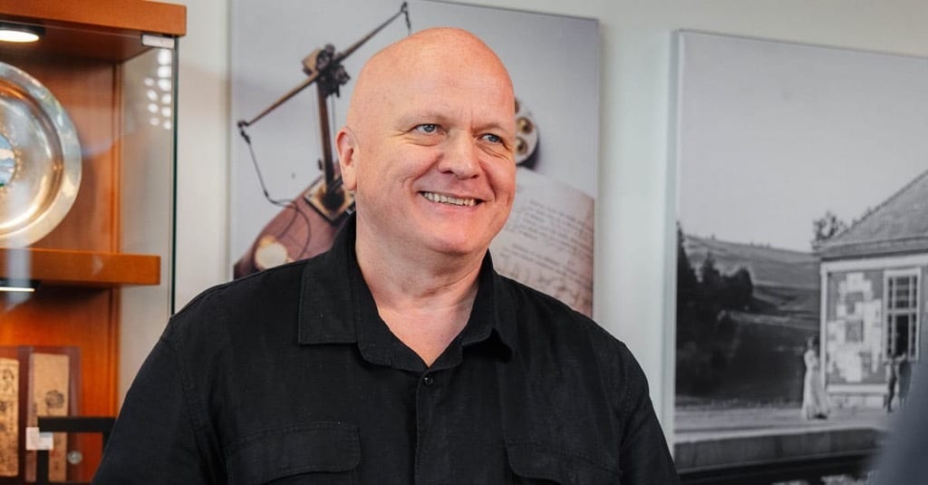 Smiling bald man in black shirt, with display cases and framed pictures behind him.
