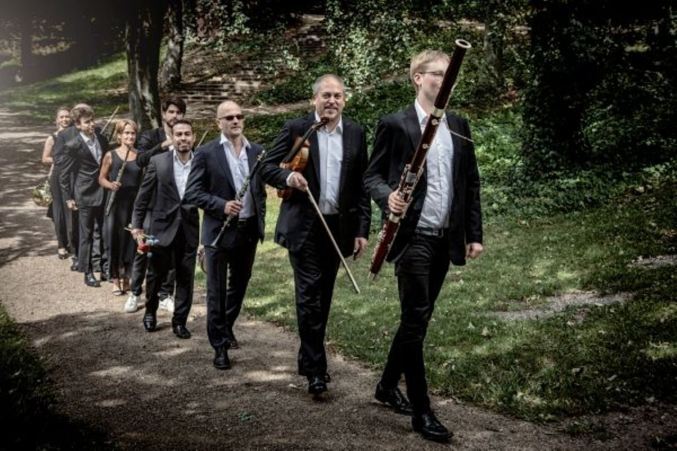 An ensemble of musicians, carrying various instruments, walks along a path in a park.