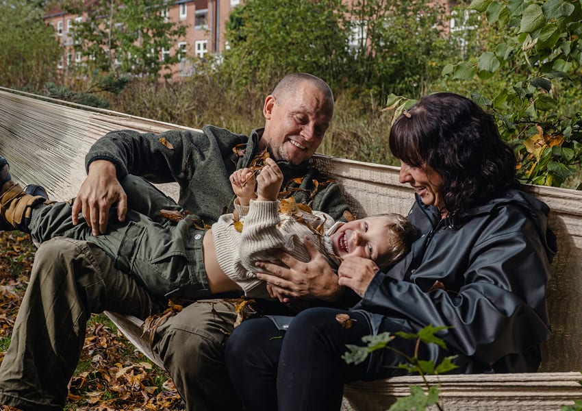 A family of three, a man, woman, and child, laughing while sitting in a hammock covered in autumn leaves.